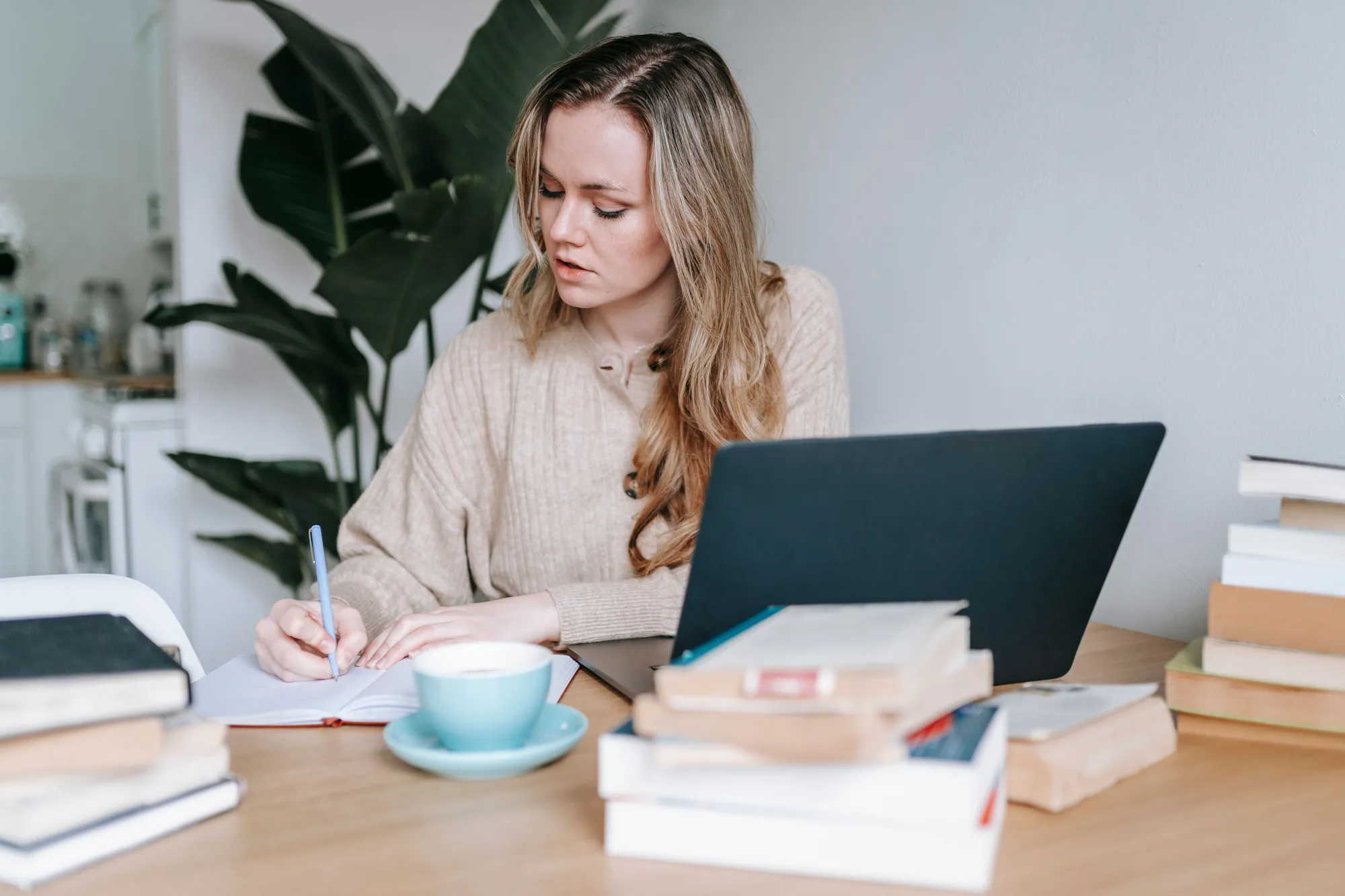 Person arbeitet in ruhiger Umgebung mit Laptop, Notizbuch und Büchern an einer Abschlussarbeit.